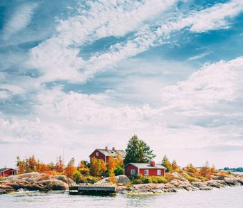 Suomi Or Finland. Beautiful Red Finnish Wooden Log Cabin House On Rocky Island Coast In Summer Sunny Evening. Lake Or River Landscape. Tiny Rocky Island Near Helsinki, Finland.